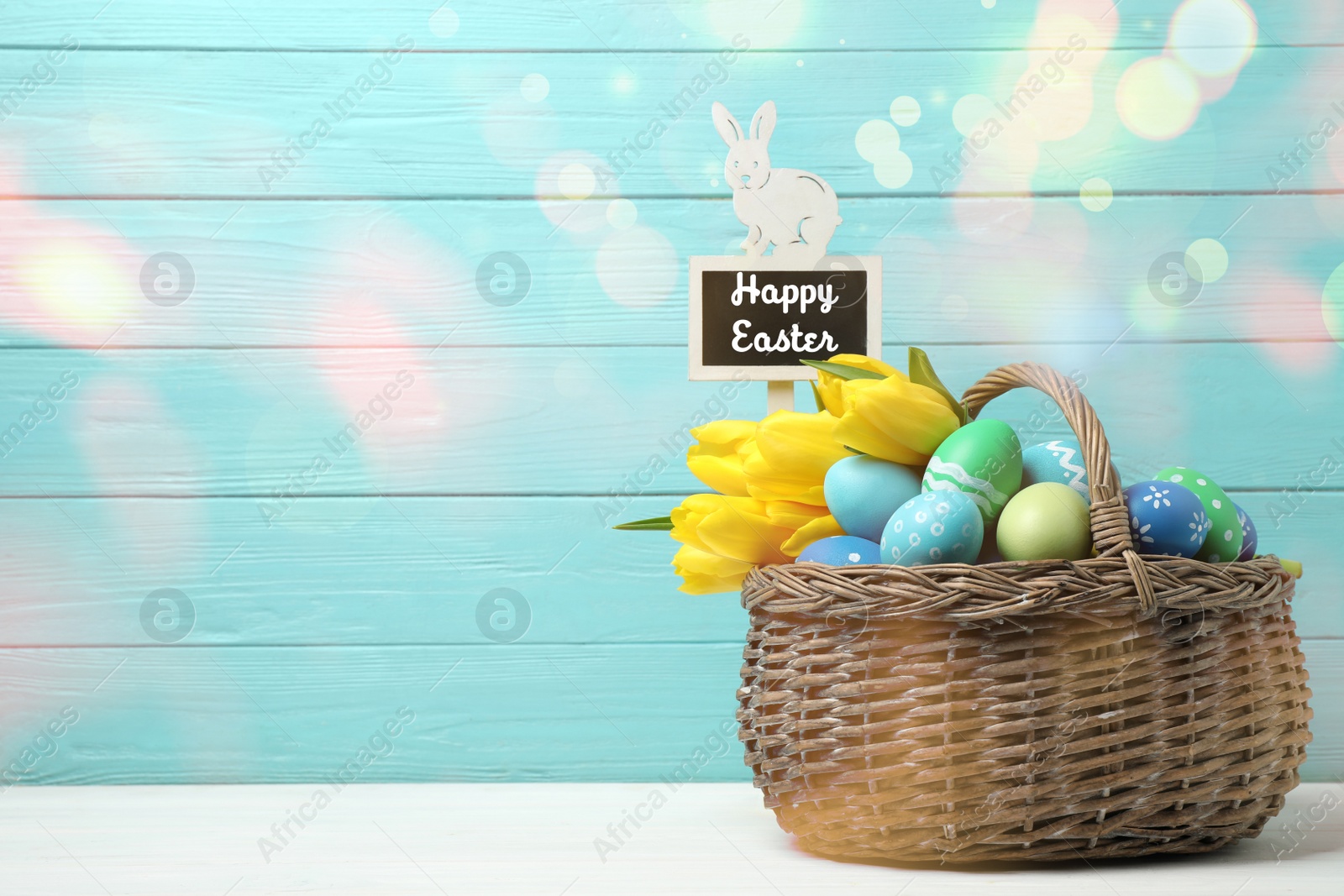 Image of Colorful eggs, tulips and tag with text Happy Easter in wicker basket on white table against light blue background