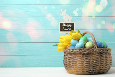 Colorful eggs, tulips and tag with text Happy Easter in wicker basket on white table against light blue background