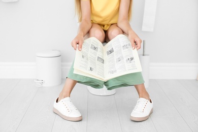 Photo of Young woman reading newspaper while sitting on toilet bowl at home