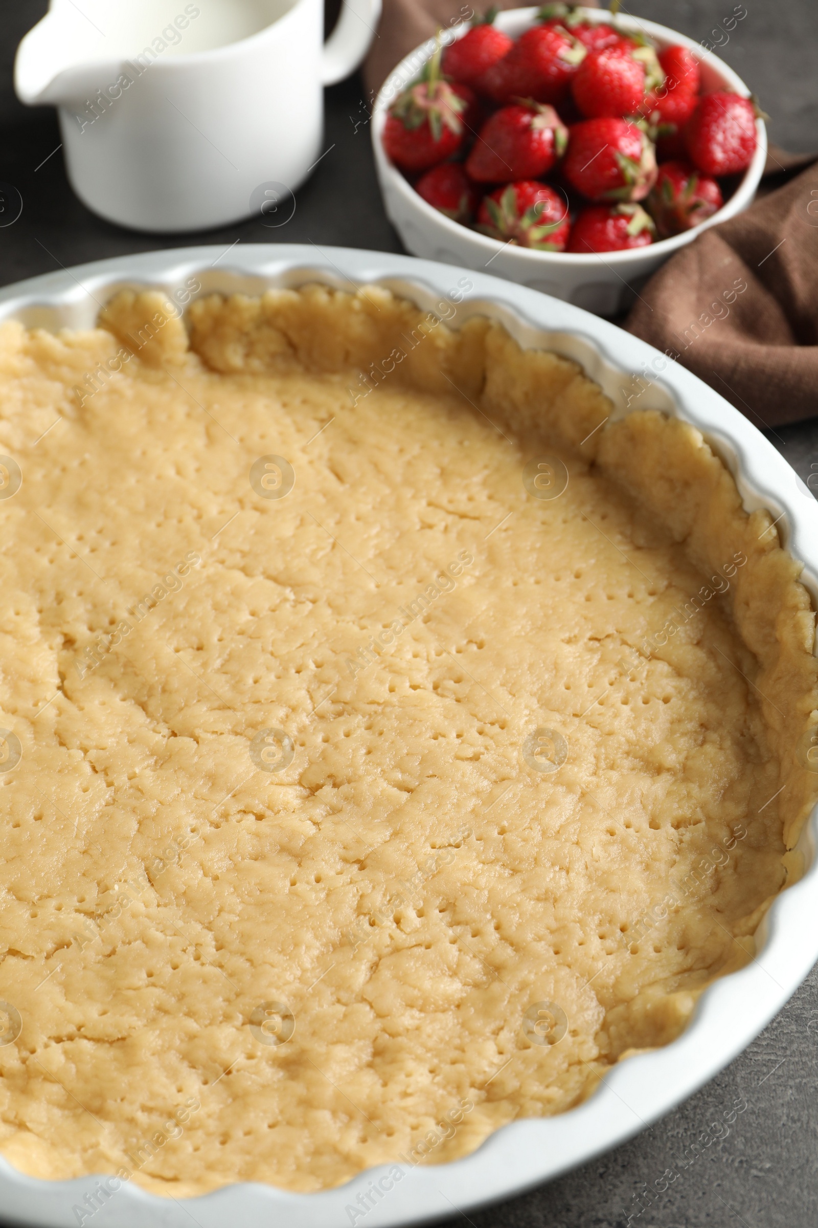 Photo of Making shortcrust pastry. Raw dough in baking dish, strawberries and milk on grey table