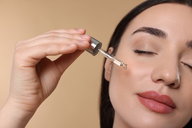 Young woman applying essential oil onto face on beige background, closeup