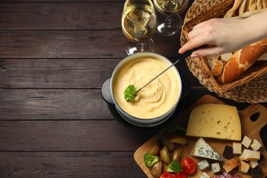 Woman dipping piece of broccoli into fondue pot with melted cheese at wooden table with wine and snacks, top view. Space for text