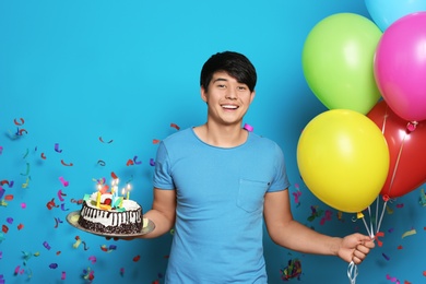 Young man with birthday cake and bright balloons on color background