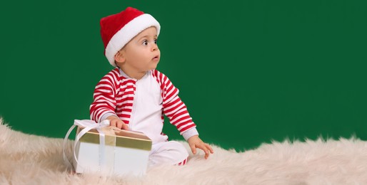 Image of Cute baby in Santa hat with Christmas gift on fluffy plaid against green background, space for text. Banner design