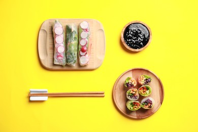 Different delicious spring rolls, chopsticks and soy sauce on yellow background, flat lay