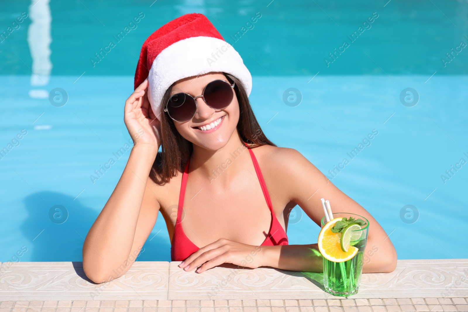 Photo of Young woman wearing Santa Claus hat with refreshing drink in swimming pool. Christmas vacation