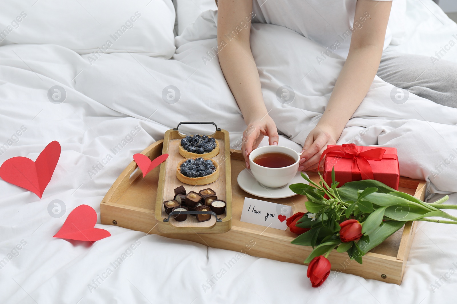 Photo of Tasty breakfast served in bed. Woman with tea, desserts, gift box, flowers and I Love You card at home, closeup