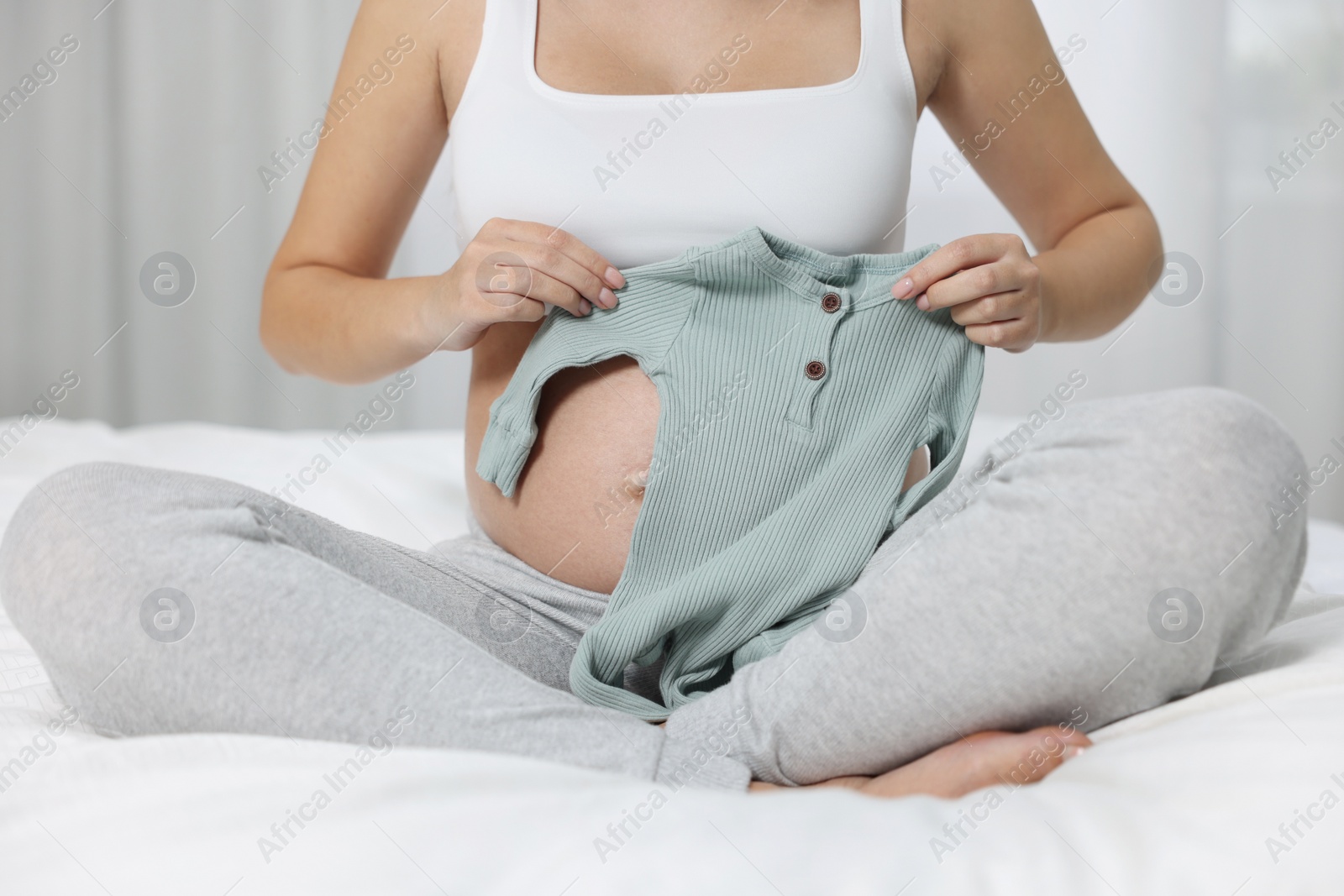 Photo of Pregnant woman with baby bodysuit sitting on bed indoors, closeup