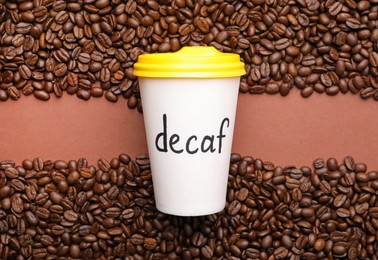 Paper cup of coffee with word Decaf and beans on brown background, flat lay