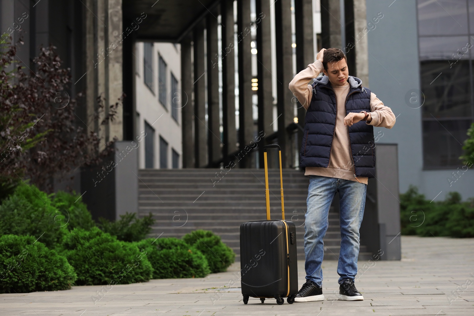 Photo of Being late. Worried man with suitcase looking at watch near building outdoors, space for text