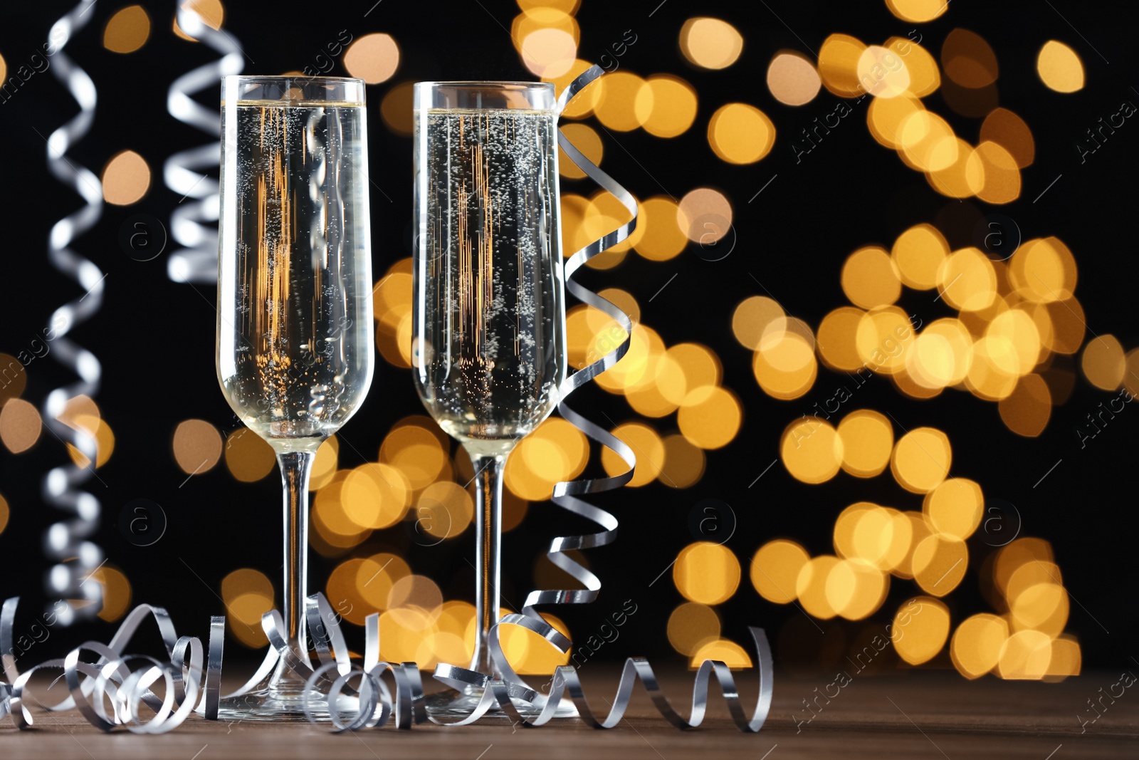 Photo of Glasses of champagne and serpentine streamers against black background with blurred lights. Space for text