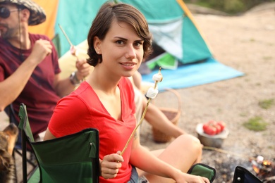 Photo of Young woman with fried marshmallows on skewer near camping tent