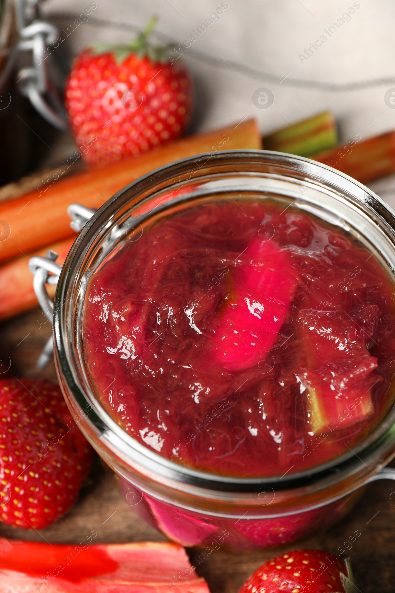 Photo of Jar of tasty rhubarb jam, fresh stems and strawberries on wooden table, closeup
