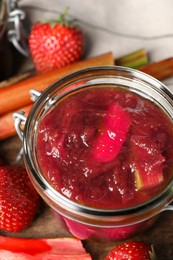 Jar of tasty rhubarb jam, fresh stems and strawberries on wooden table, closeup
