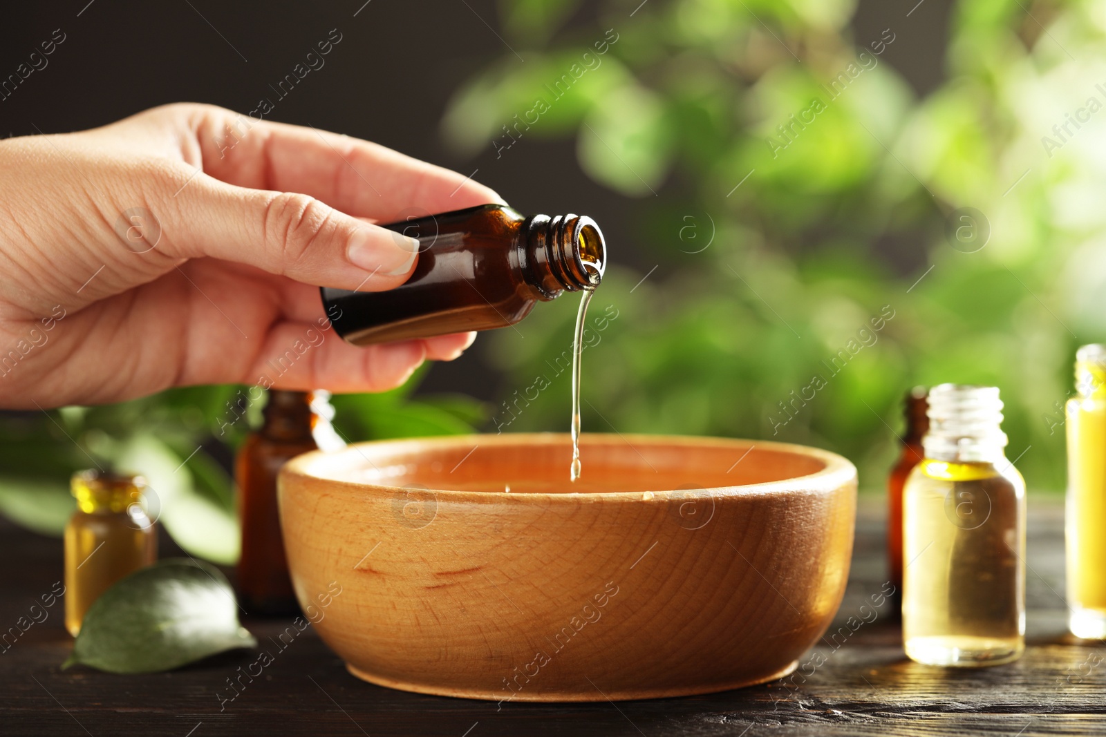 Photo of Woman pouring essential oil from glass bottle into bowl on table, closeup