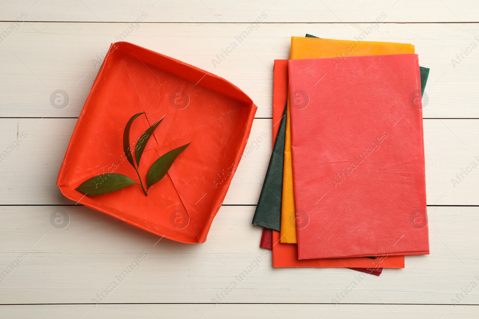 Photo of Colorful beeswax food wraps and fresh leaves on white wooden table, flat lay