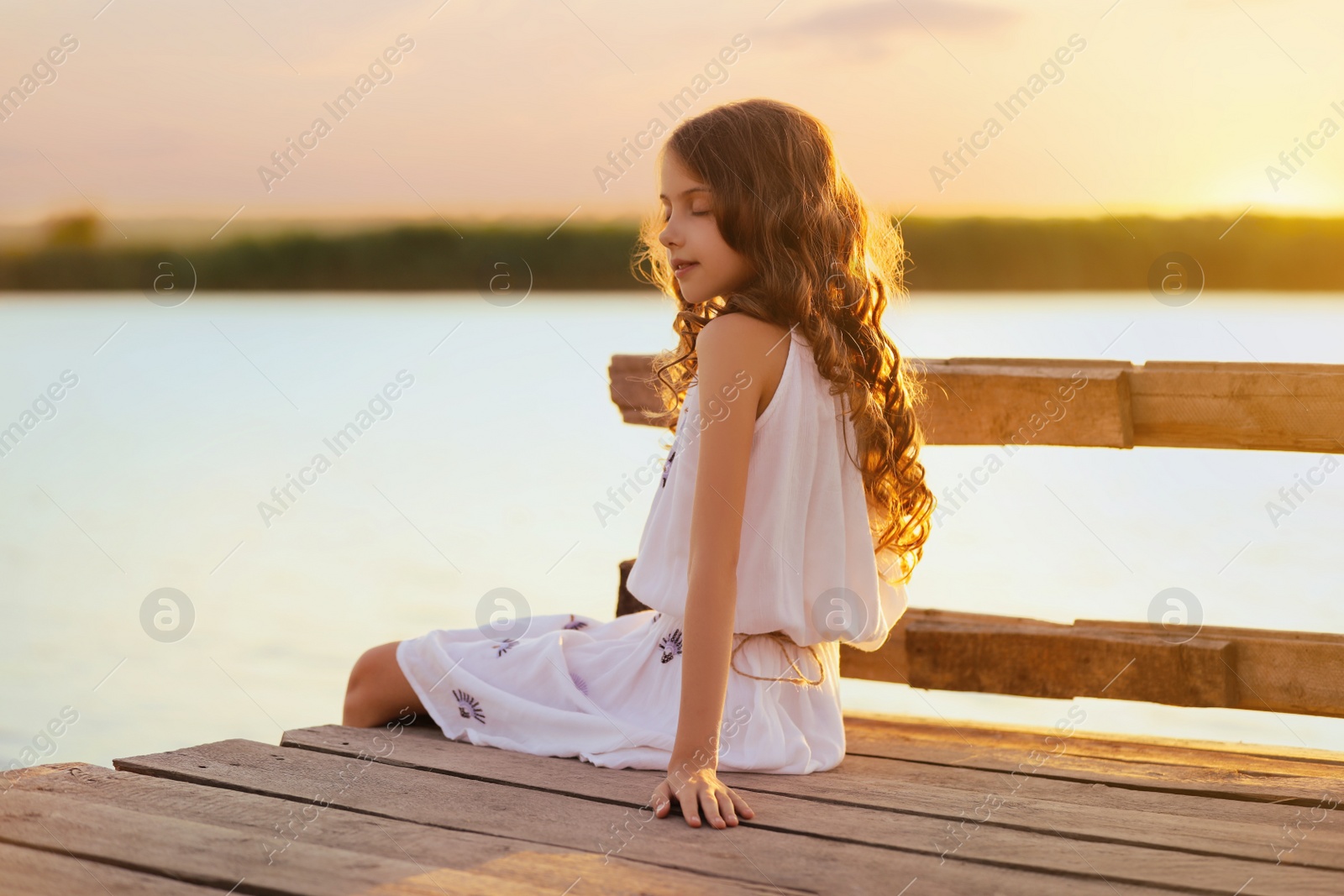 Photo of Cute little girl sitting on wooden pier near river on sunny day