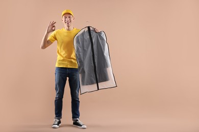 Dry-cleaning delivery. Happy courier holding garment cover with clothes and showing OK gesture on beige background, space for text