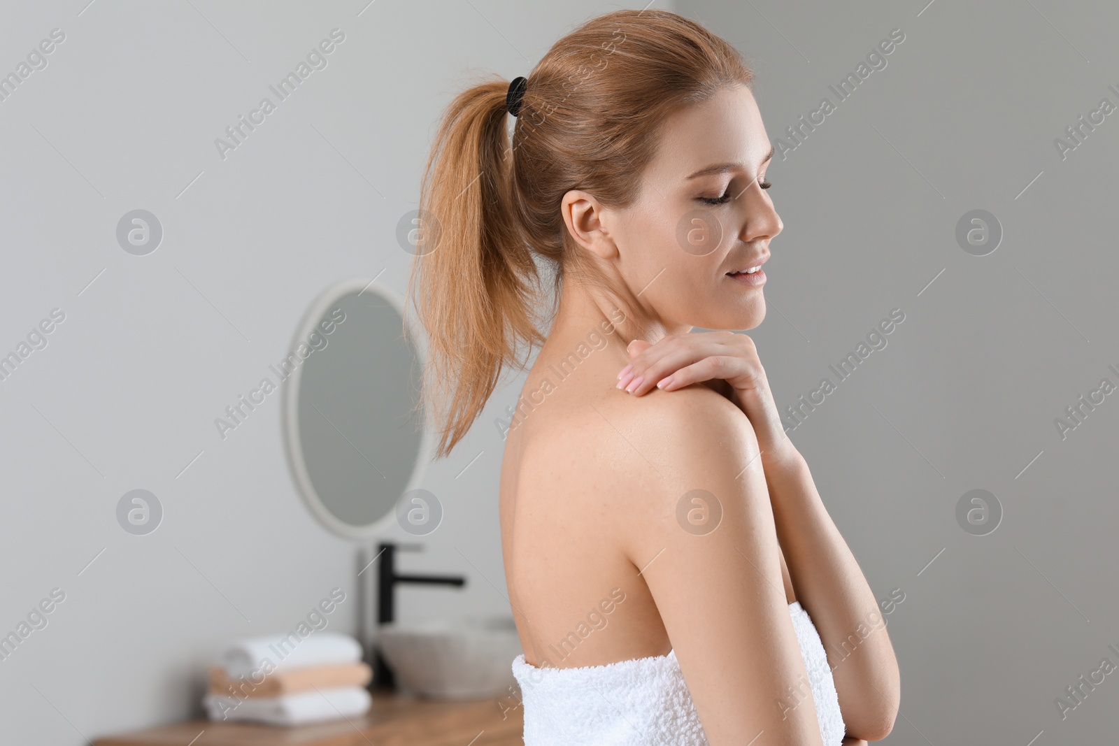 Photo of Beautiful woman applying body oil onto shoulder in bathroom, space for text