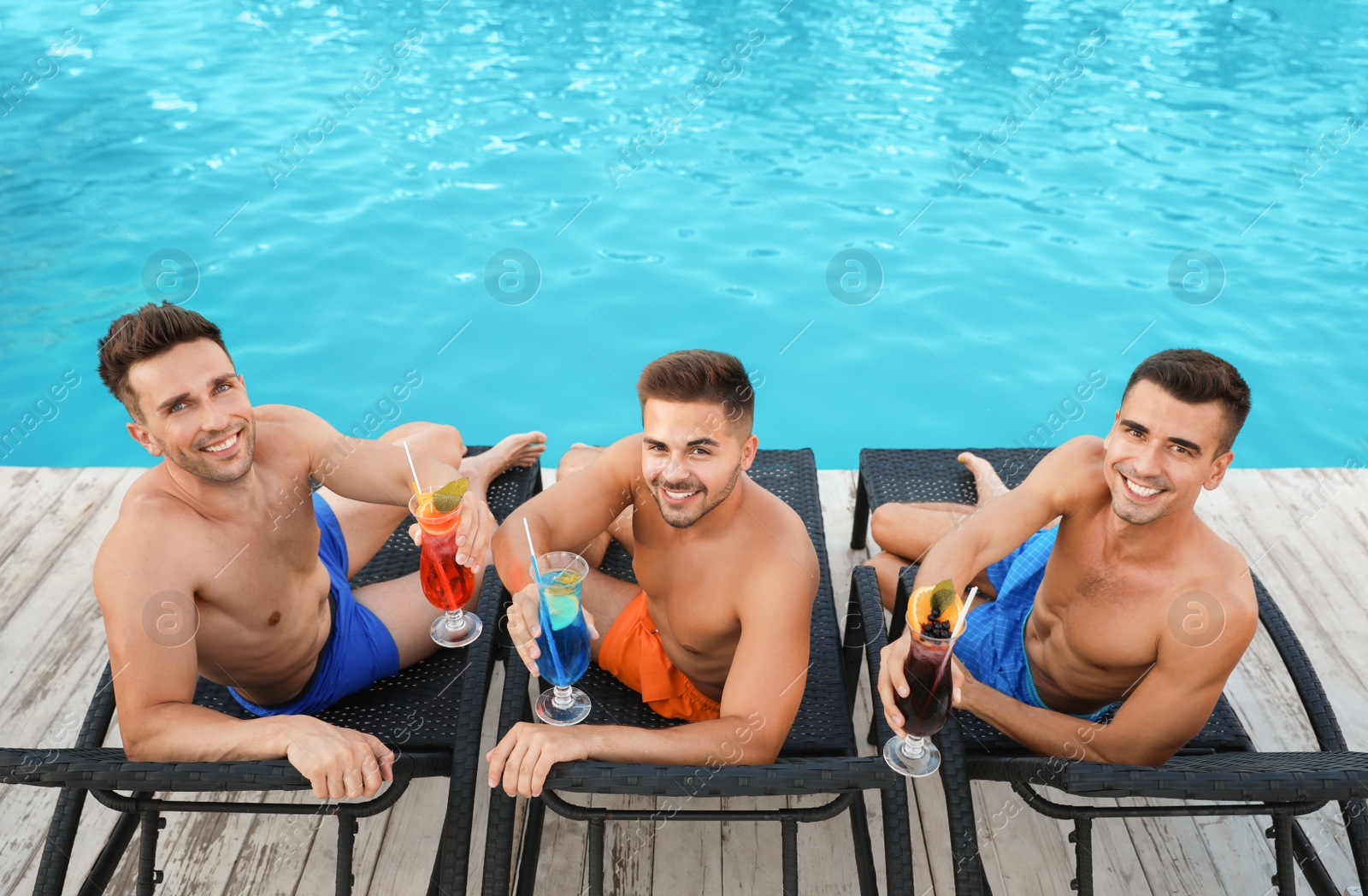 Photo of Happy young friends with refreshing cocktails relaxing on deck chairs near swimming pool