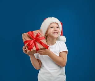 Photo of Cute little child wearing Santa hat with Christmas gift on blue background