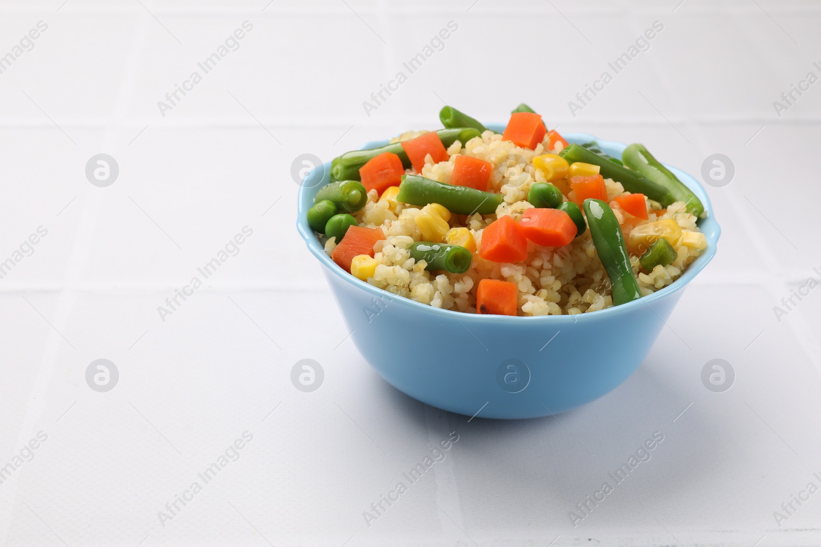 Photo of Delicious bulgur with vegetables in bowl on white tiled table, closeup. Space for text