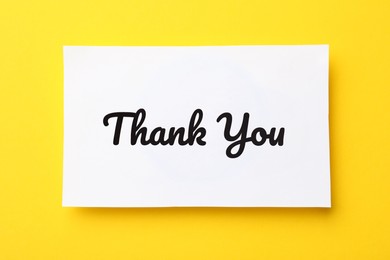 Photo of White paper note with phrase Thank You on yellow background, top view