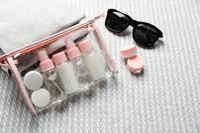 Photo of Cosmetic travel kit. Plastic bag with small containers of personal care products, stack of clothes and sunglasses on sofa