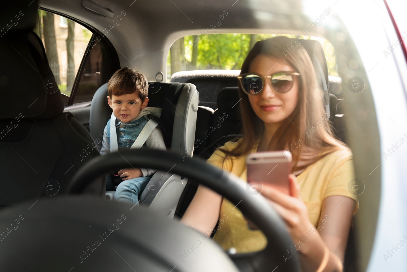 Photo of Mother using phone while driving car with her son on backseat. Child in danger
