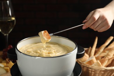 Woman dipping piece of bread into fondue pot with melted cheese at table, closeup