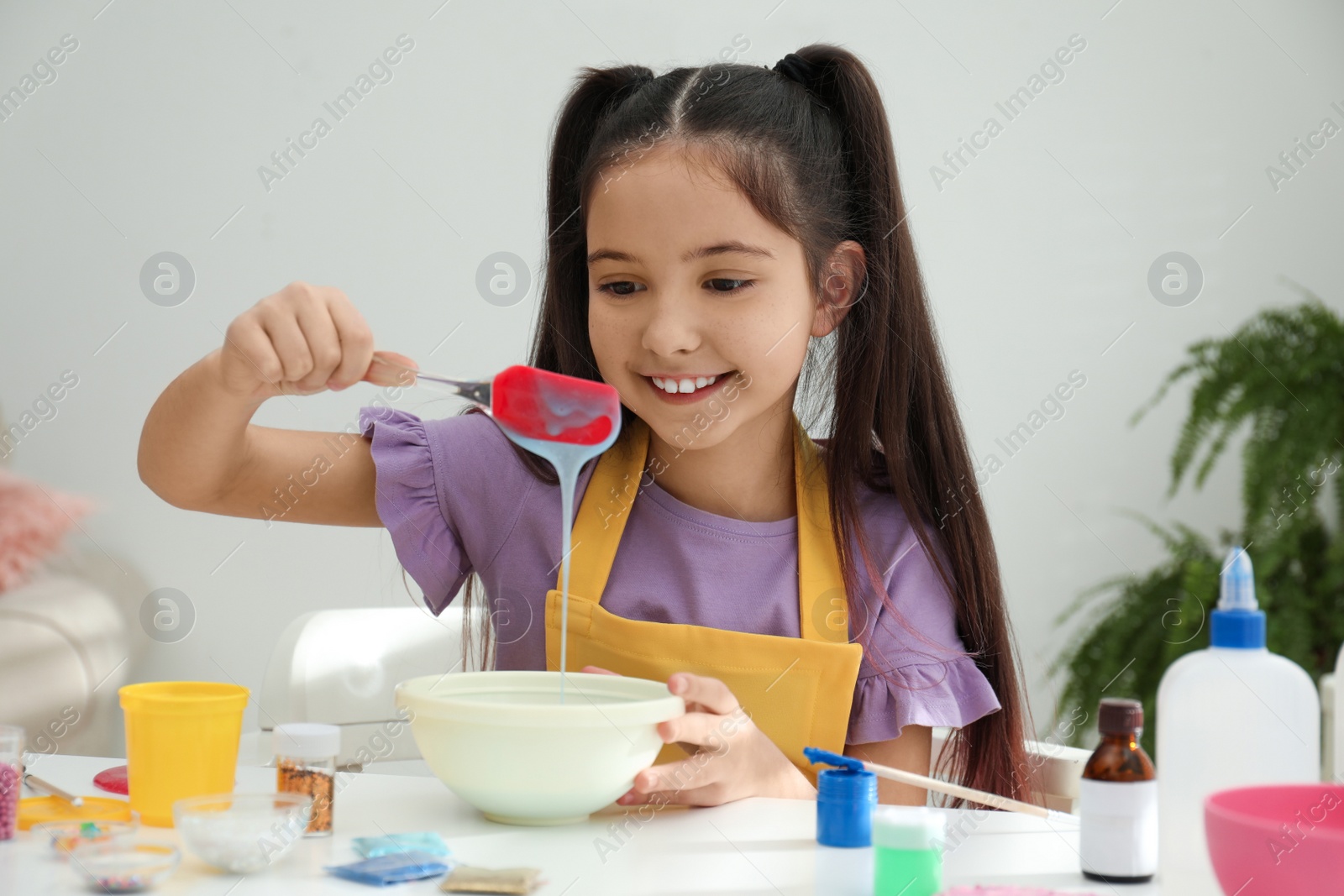 Photo of Cute little girl mixing ingredients with silicone spatula at table in room. DIY slime toy