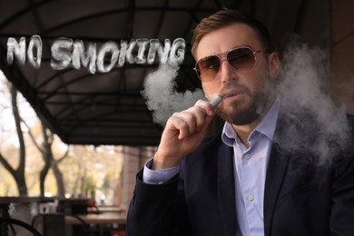 Image of Phrase No Smoking of smoke near businessman using electronic cigarette at table in outdoor cafe
