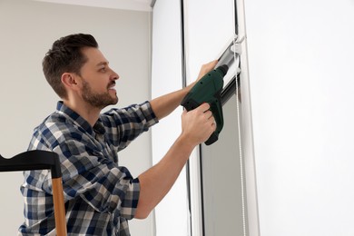 Photo of Man with drill installing roller window blind on stepladder indoors