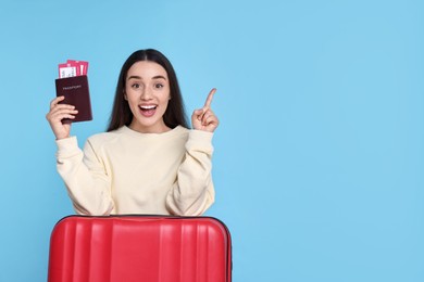 Happy woman with passport, tickets and suitcase pointing at something on light blue background. Space for text