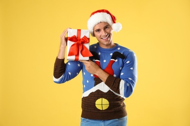 Photo of Happy man in Christmas sweater and Santa hat holding gift box on yellow background