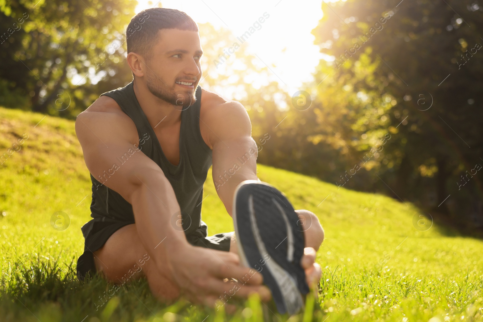 Photo of Attractive man doing exercises on green grass in park, space for text. Stretching outdoors