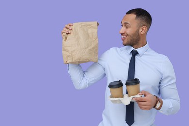 Photo of Happy young intern holding takeaway cups with hot drink and paper bag on lilac background. Space for text
