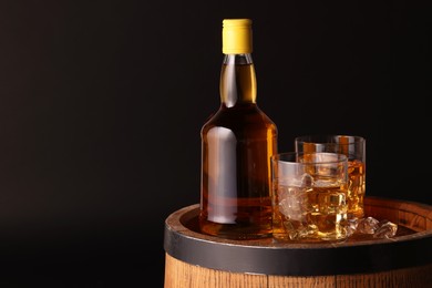 Whiskey with ice cubes in glasses and bottle on wooden barrel against black background, space for text
