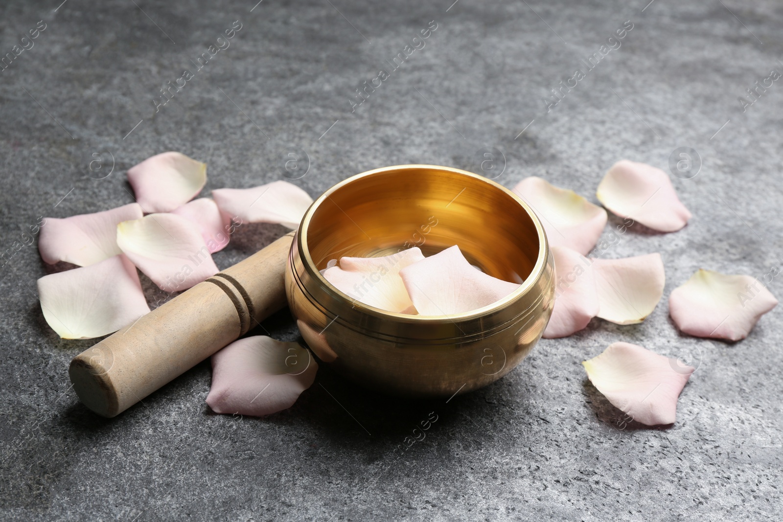 Photo of Golden singing bowl with petals and mallet on grey table. Healing instrument