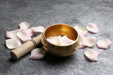 Photo of Golden singing bowl with petals and mallet on grey table. Healing instrument