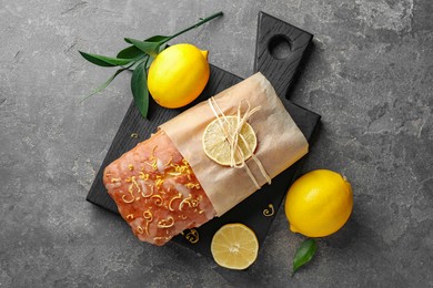 Photo of Wrapped tasty lemon cake with glaze and citrus fruits on grey textured table, flat lay