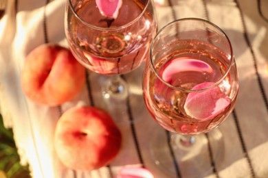 Photo of Glasses of delicious rose wine with petals and peaches on white picnic blanket, above view