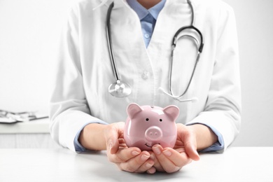 Photo of Doctor with piggybank at white table indoors, closeup. Medical insurance concept