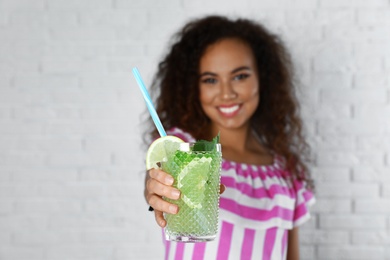Photo of Happy African-American woman with glass of natural lemonade near white brick wall, closeup. Detox drink