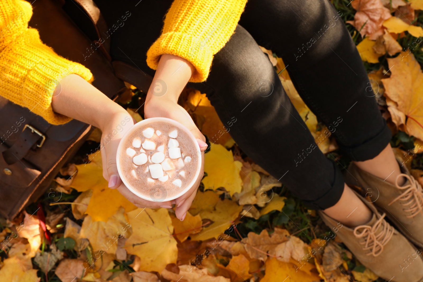 Photo of Woman holding cup of hot drink in park with fallen leaves, above view. Autumn season