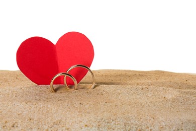 Photo of Honeymoon concept. Two golden rings, red paper heart and sand isolated on white