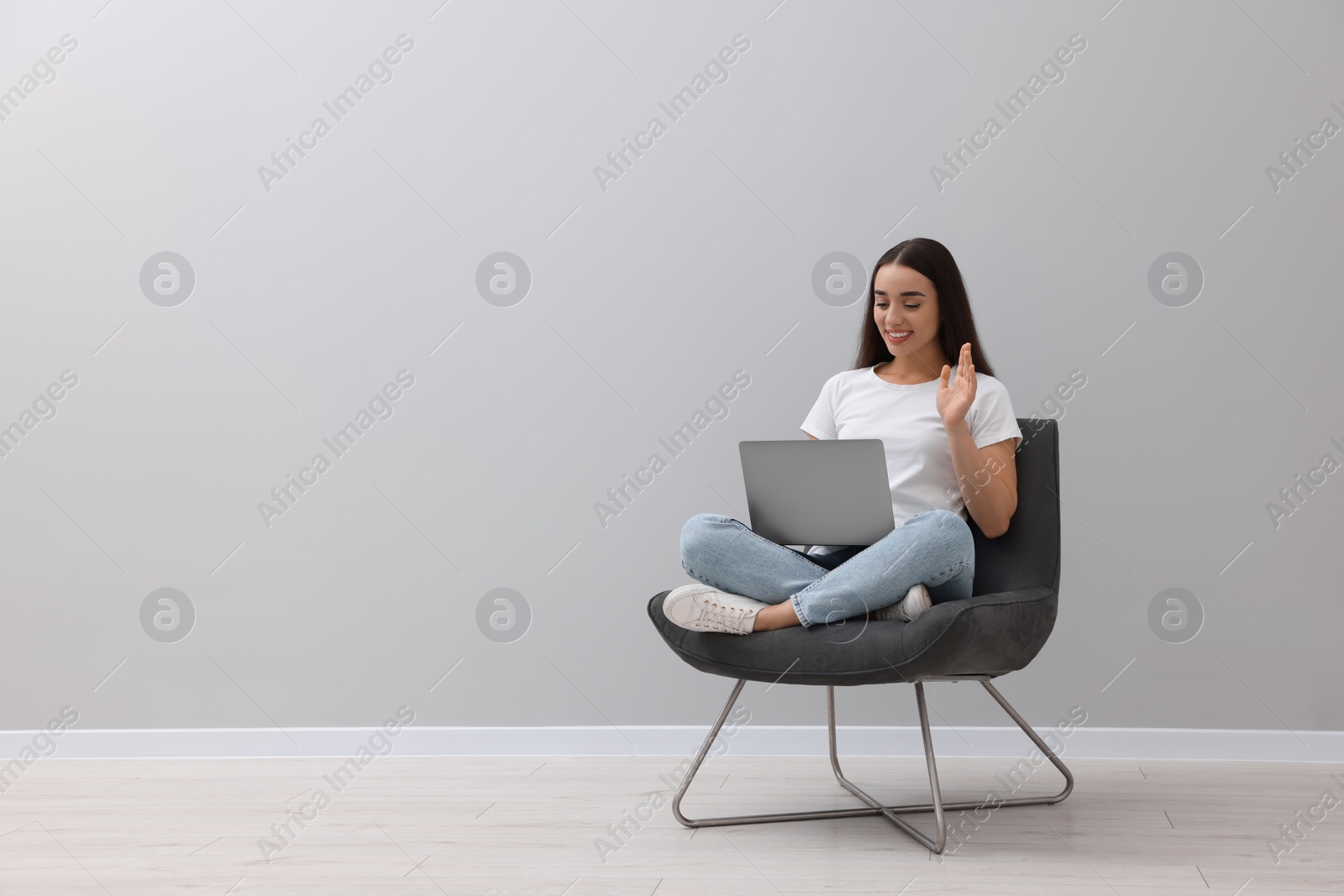 Photo of Beautiful woman having video chat via laptop while sitting in armchair near light grey wall indoors, space for text