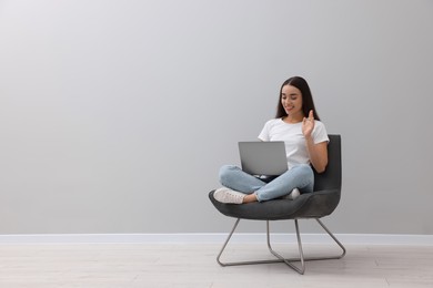 Beautiful woman having video chat via laptop while sitting in armchair near light grey wall indoors, space for text