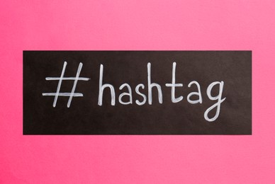 Black paper with word Hashtag and symbol on pink background, top view