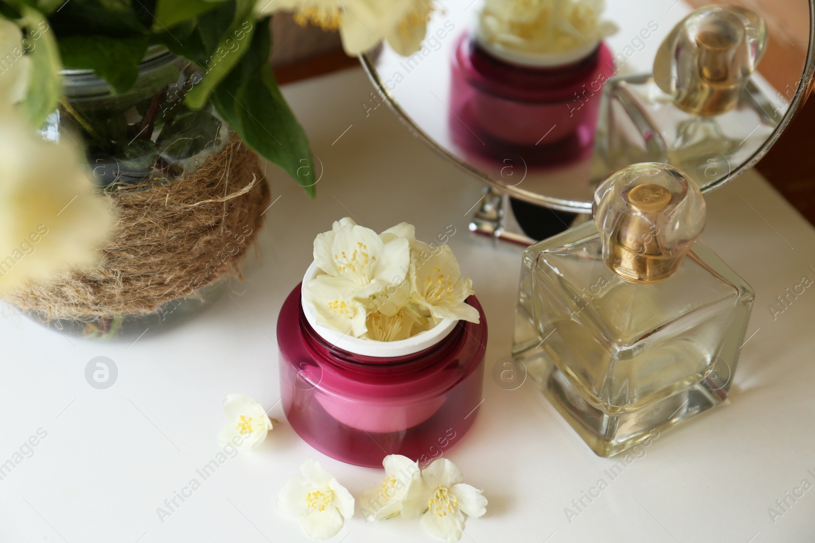 Photo of Jar of cream with beautiful jasmine flowers, perfume and mirror on white table